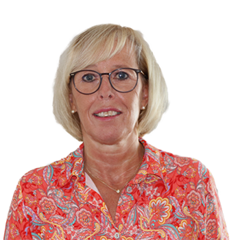 Frau mit blonden, kurzen Haaren, Brille und bunter Bluse mit Blumenmuster – freundlich lächelnd vor neutralem Hintergrund.