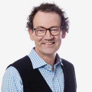 Das Bild zeigt einen Mann mit lockigem, dunklem Haar und Brille. Er trägt ein hellblau-kariertes Hemd und eine schwarze Weste. Der Hintergrund ist weiß, und er lächelt freundlich in die Kamera.